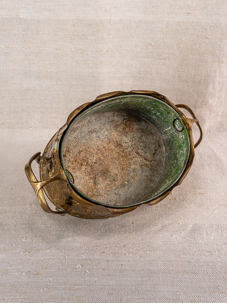 Vintage Swiss brass jardinière with embossed fruit motif, oval shape, and greenish interior on beige fabric background.