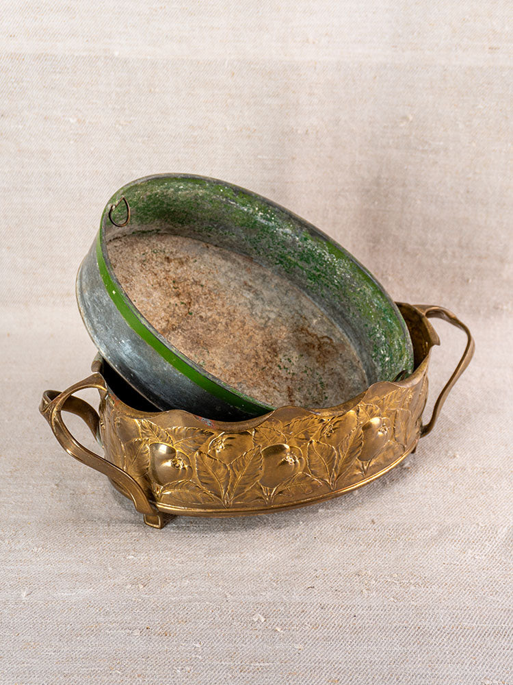 Vintage Swiss brass jardinière with handles and embossed fruit motifs, green metal bowl inside, beige fabric backdrop.