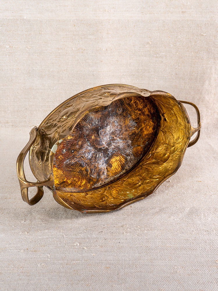 Vintage Swiss brass jardiniere with embossed fruit motif, twin handles, and tarnished interior on woven fabric background.