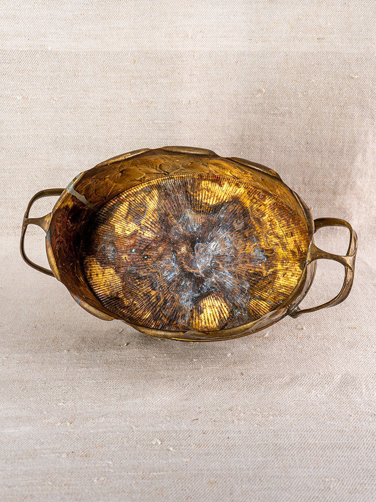 Vintage Swiss brass jardinière with embossed fruit motif and side handles, multicolored interior, beige fabric backdrop.