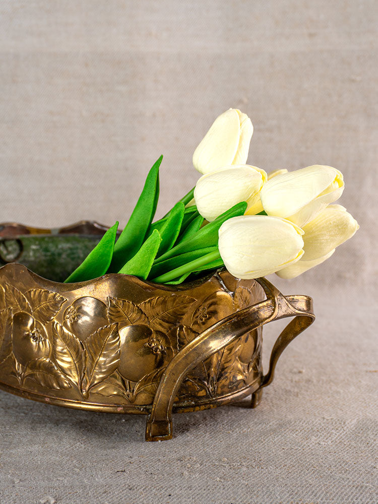 Vintage Swiss brass jardinière with embossed fruit motif, holding pale yellow tulips, on a neutral fabric background.
