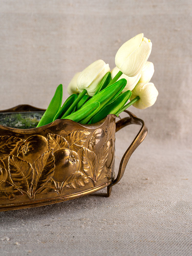 Vintage Swiss brass jardinière with embossed fruit and two handles, green glass liner, set on neutral fabric.