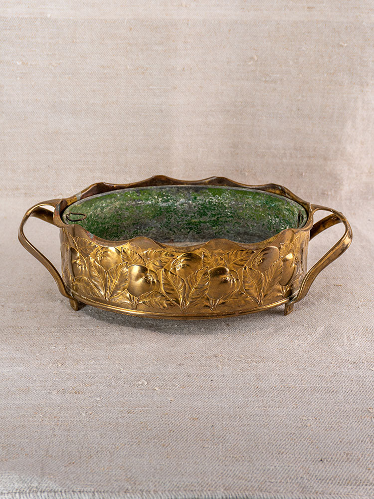 Vintage Swiss brass jardinière with embossed fruit and two handles, green glass liner, set on neutral fabric.