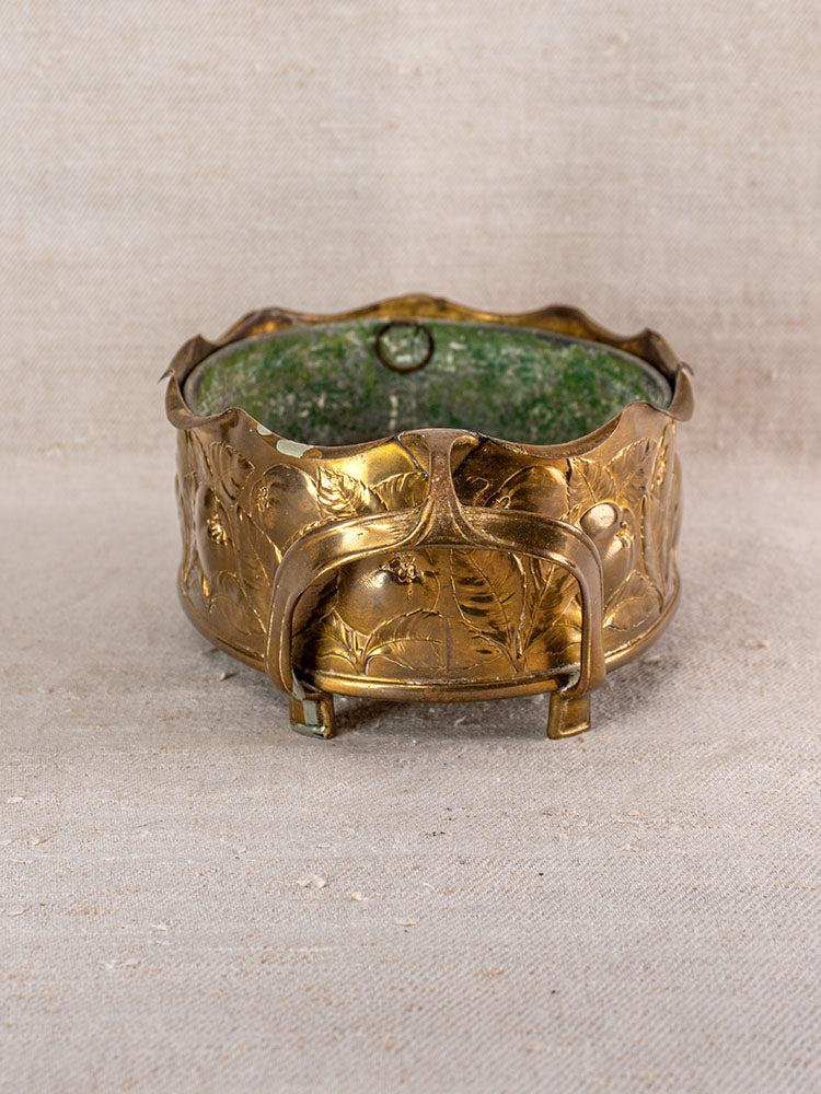 Vintage Swiss brass jardinière with embossed fruit motif, decorative handles, and green glass liner on neutral fabric.