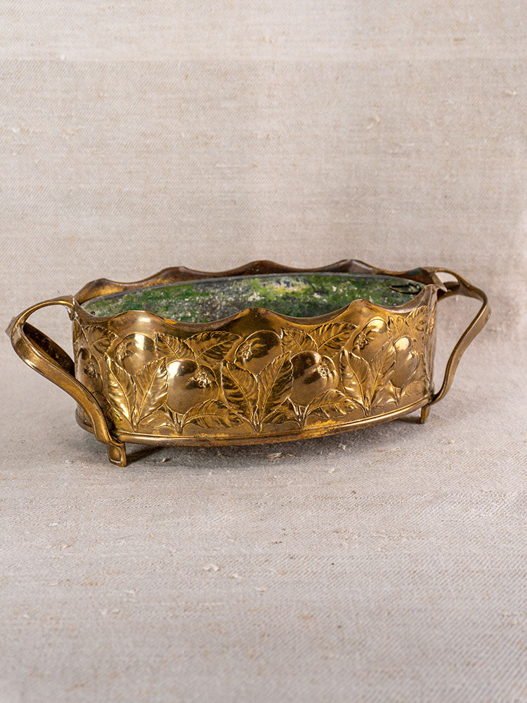 Vintage Swiss brass jardinière with embossed fruit, two handles, wavy rim, green patina, on beige textured fabric.