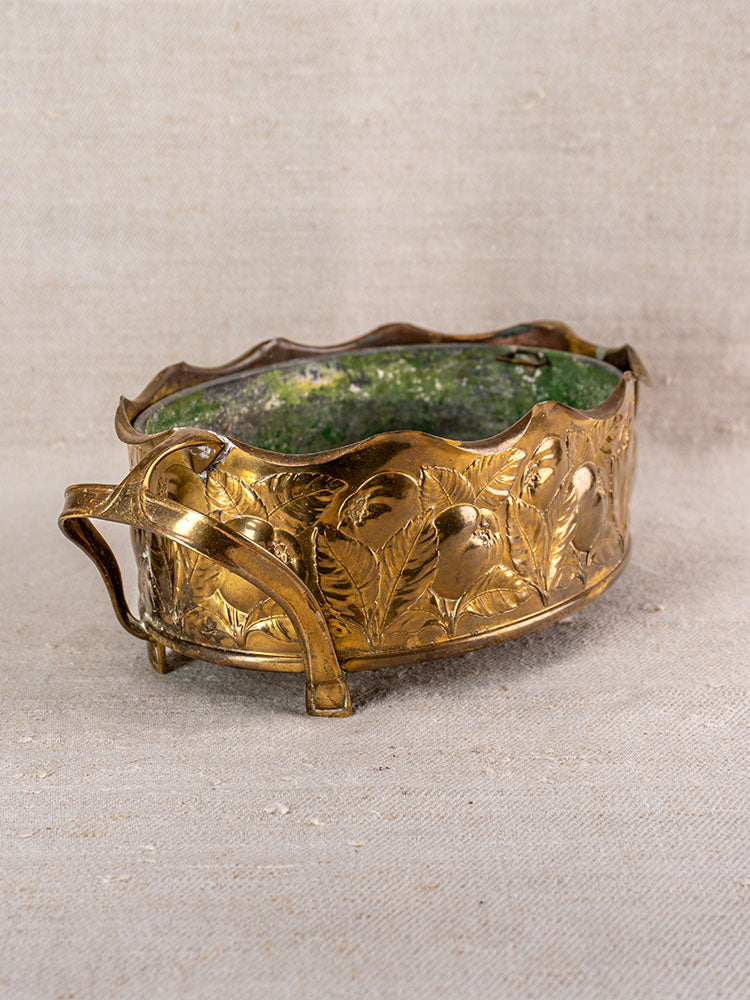 Vintage Swiss brass jardinière with embossed fruit, curved handles, scalloped rim, and green interior on beige fabric.
