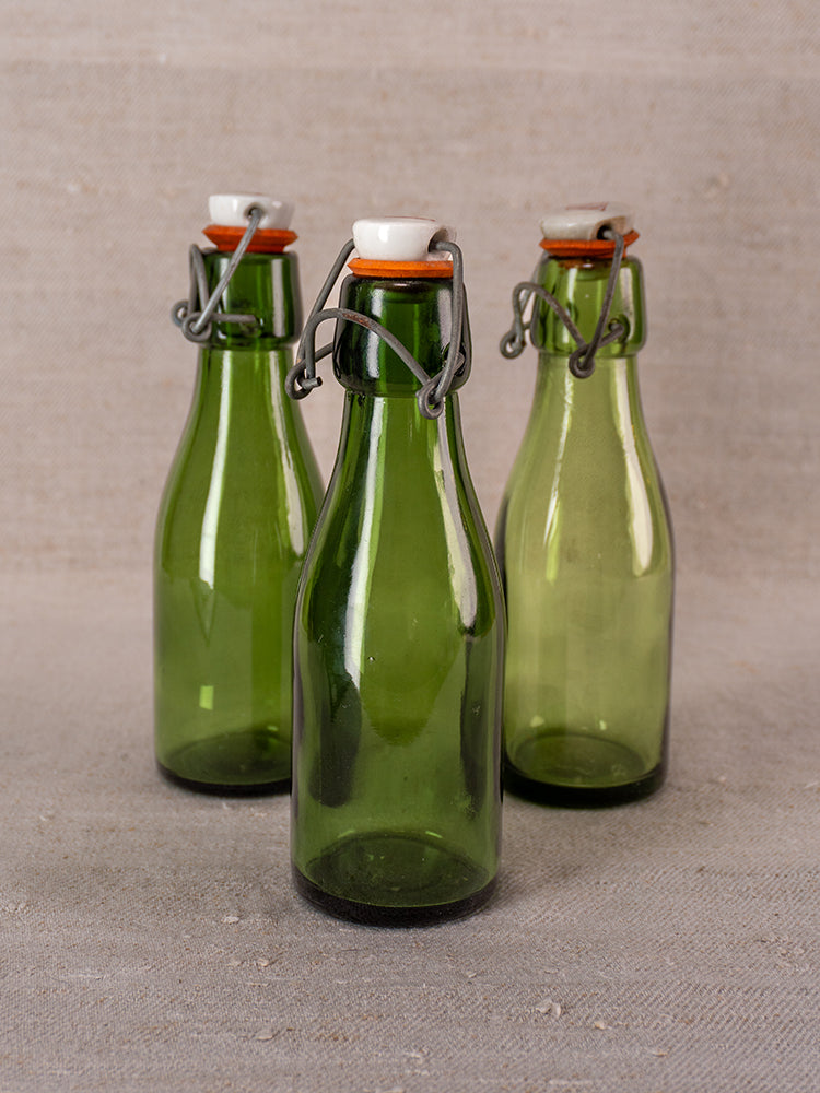 Three Swiss Bülach green glass swing-top bottles with porcelain tops displayed on beige fabric background.
