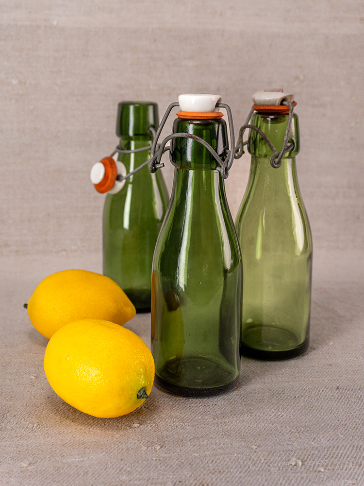 Set of three Swiss Bülach green glass clamp bottles with porcelain tops, displayed on beige fabric with two yellow lemons.