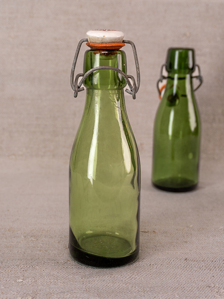 Swiss Bülach green glass clamp bottles with porcelain tops displayed on a gray surface, one in sharp focus.