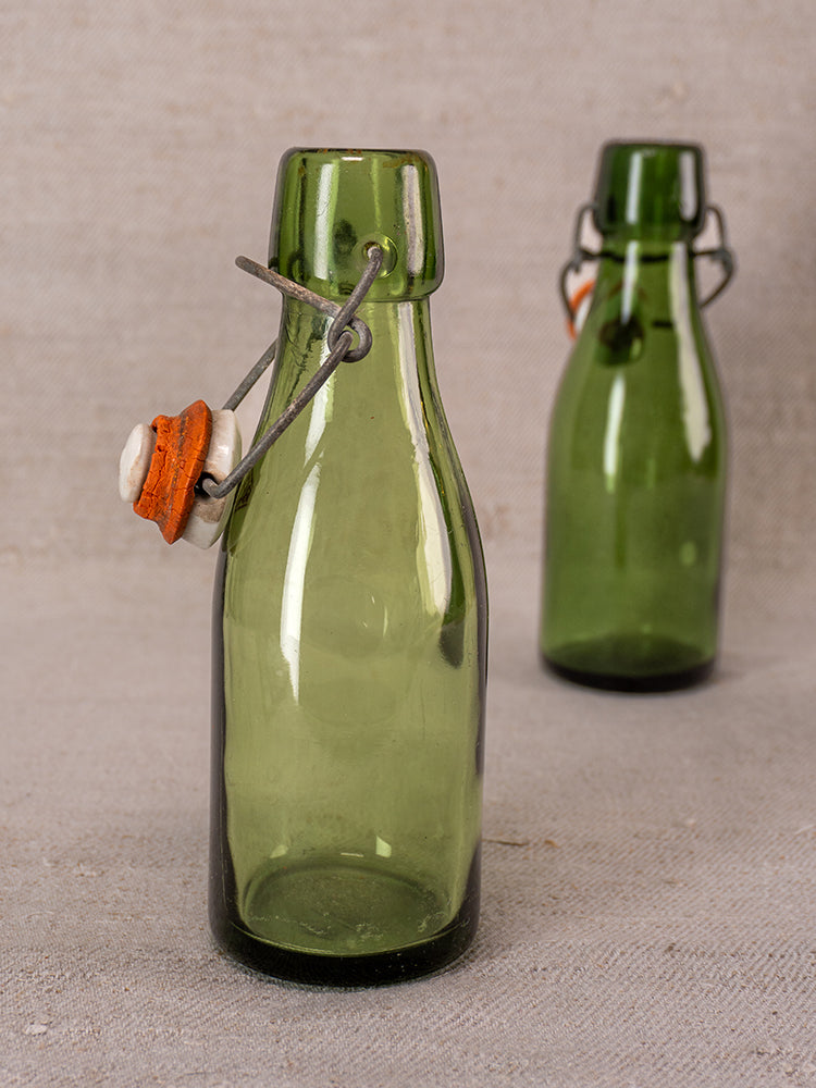 Swiss Bülach green glass clamp bottles with porcelain tops, one in focus, others softly blurred on neutral fabric.