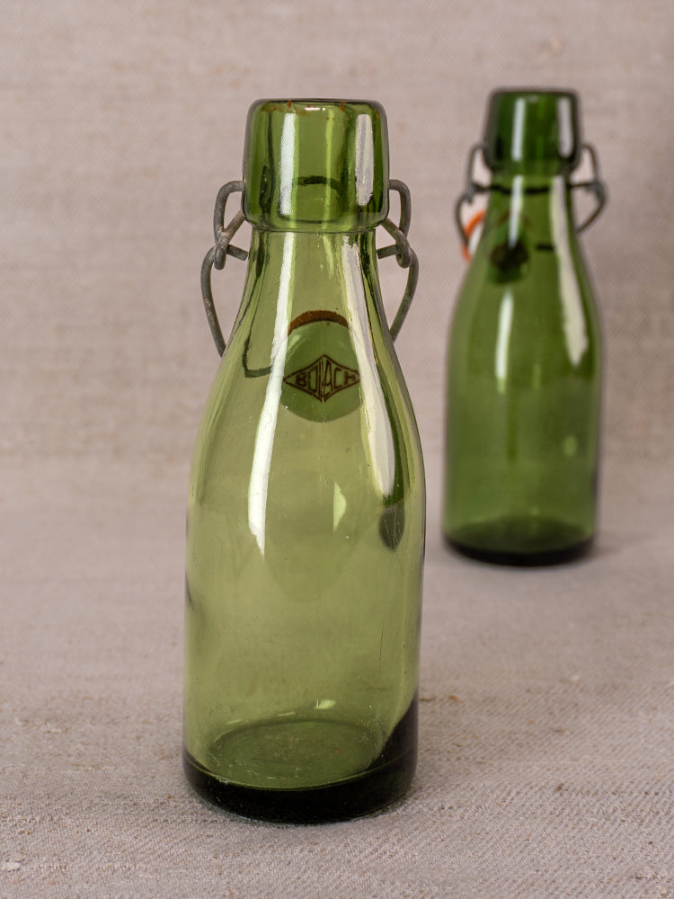 Swiss Bülach green glass clamp bottles with porcelain tops on a neutral textured surface, foreground bottle in focus.