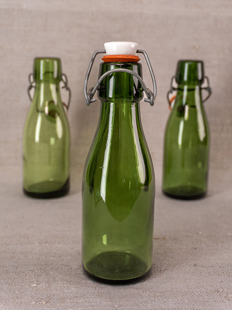 Three Swiss Bülach green glass clamp bottles with porcelain tops on a neutral textured surface, front bottle in sharp focus.