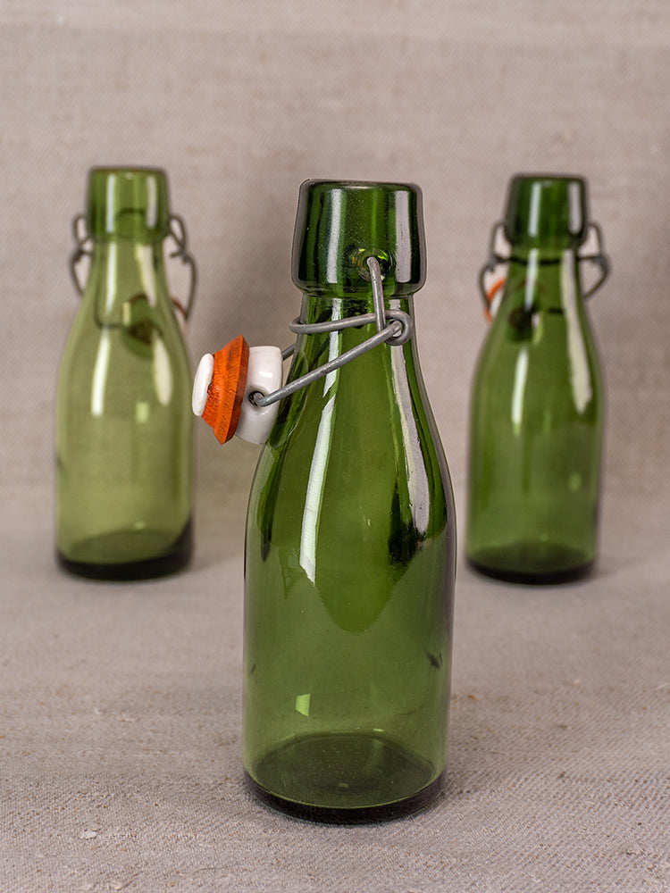 Three Swiss Bülach green glass clamp bottles with porcelain tops arranged on a neutral fabric surface. Classic collectible décor.