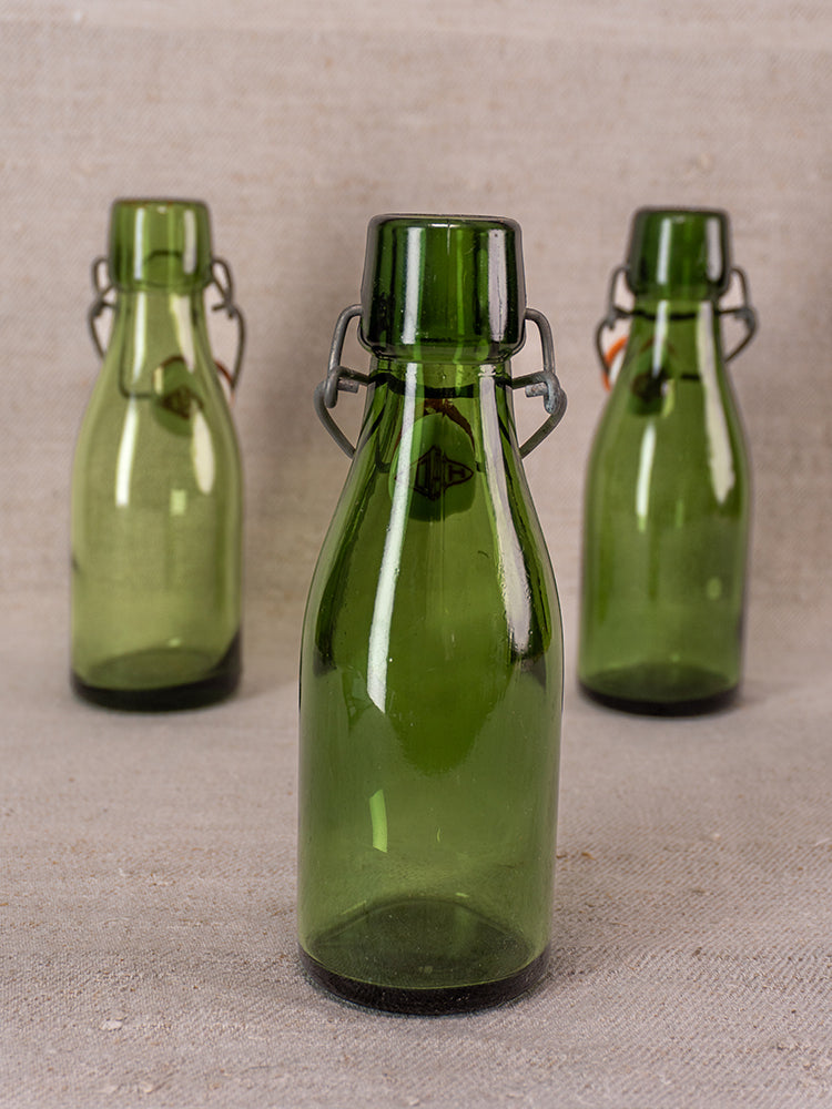 Three Swiss Bülach green glass clamp bottles with porcelain tops on a beige textured surface, foreground bottle in focus.