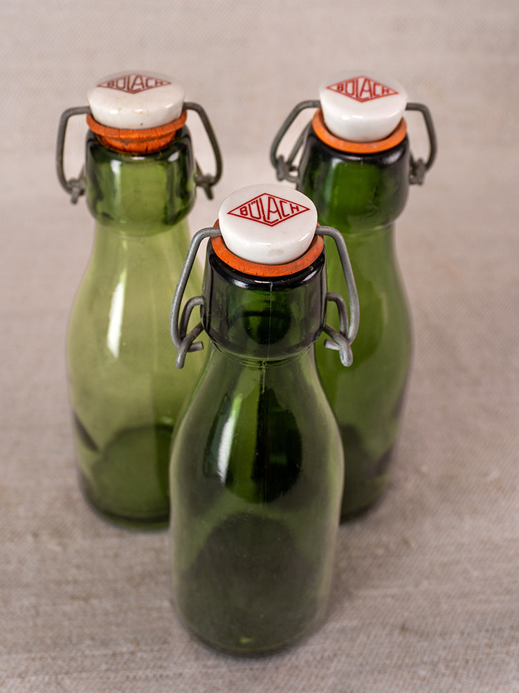 Set of three Swiss Bülach green glass clamp bottles with porcelain tops and red BOLACHA logo on light fabric.