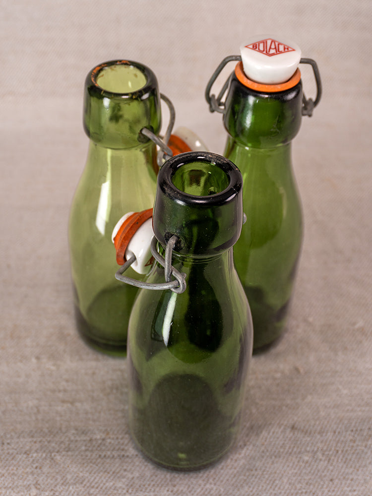 Set of three Swiss Bülach green glass clamp bottles with porcelain tops on a textured beige surface.