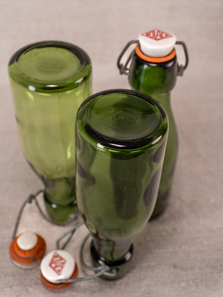Set of three Swiss Bülach green glass clamp bottles with vintage porcelain tops, on textured beige background.