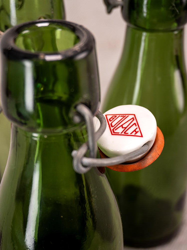 Swiss Bülach green glass clamp bottles with porcelain tops, one displaying a red geometric logo, blurred bottles behind.