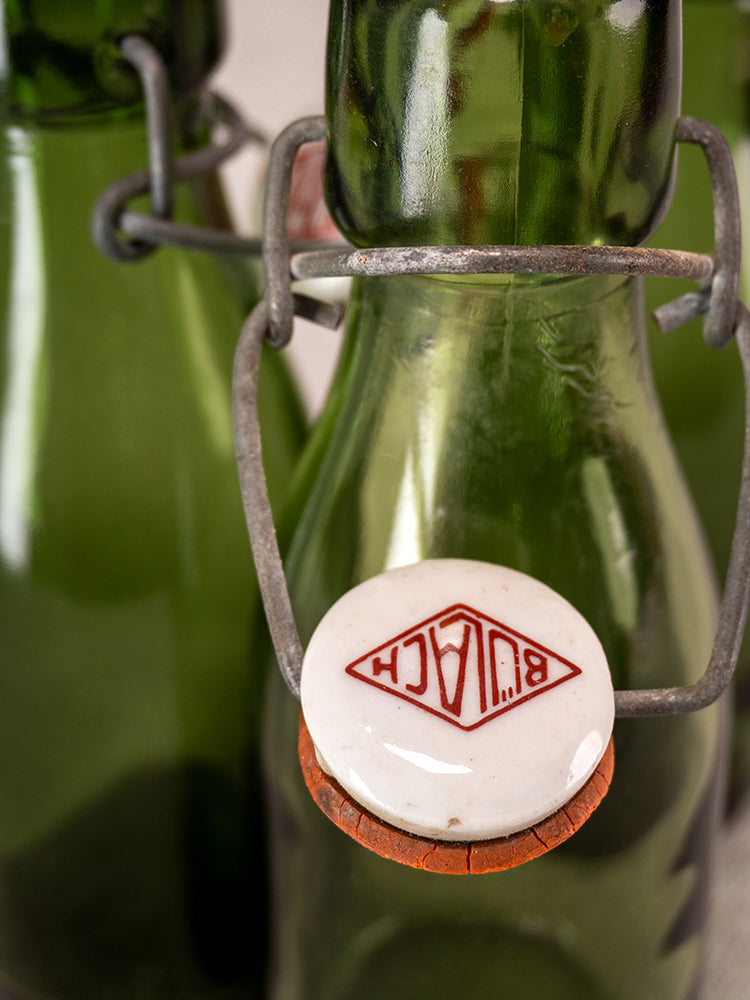 Swiss Bülach green glass clamp bottles with porcelain tops, front bottle bears red mirrored “BLAH” logo, blurred bottles behind.
