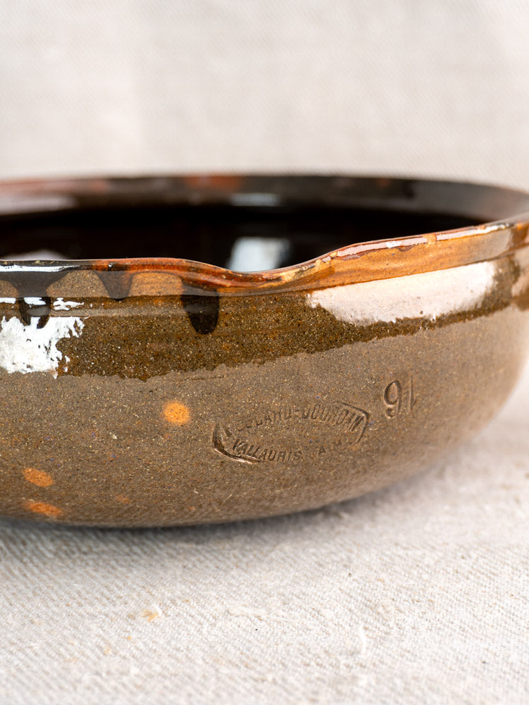Antique French Grandjean Jourdan Vallauris poêlon with rustic brown glaze, wavy rim, maker’s mark, on textured fabric.