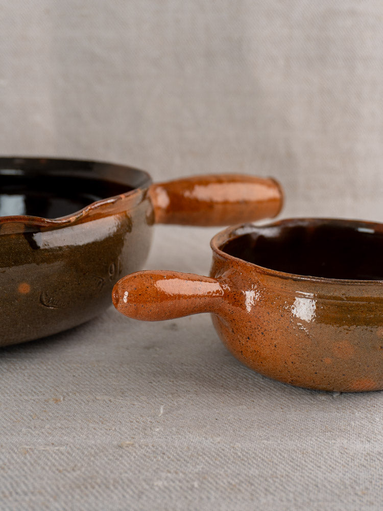 Pair of Grandjean Jourdan Vallauris French ceramic poêlon bowls in brown and orange glaze with handles on beige fabric.