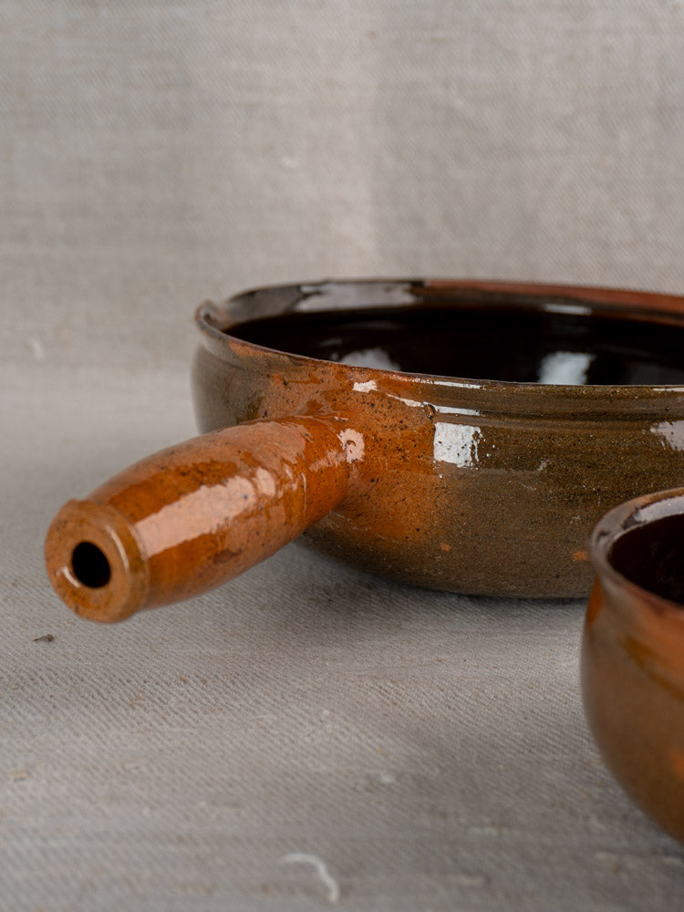 Mid-20th century French Grandjean Jourdan Vallauris brown glazed poêlon with handle, on neutral fabric. Matching bowl partially visible.