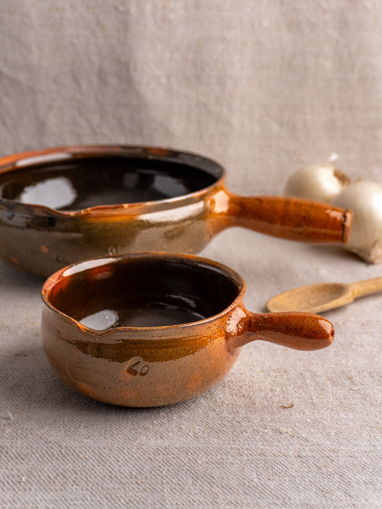 Pair of brown antique French Poêlon pans by Grandjean Jourdan Vallauris on beige cloth with wooden spoon and garlic nearby.