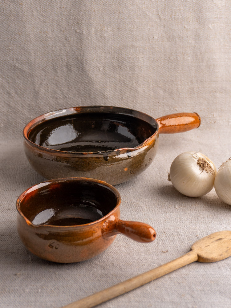 Antique French poêlon pair by Grandjean Jourdan Vallauris, brown ceramic pans on beige fabric with onions and wooden spoon.