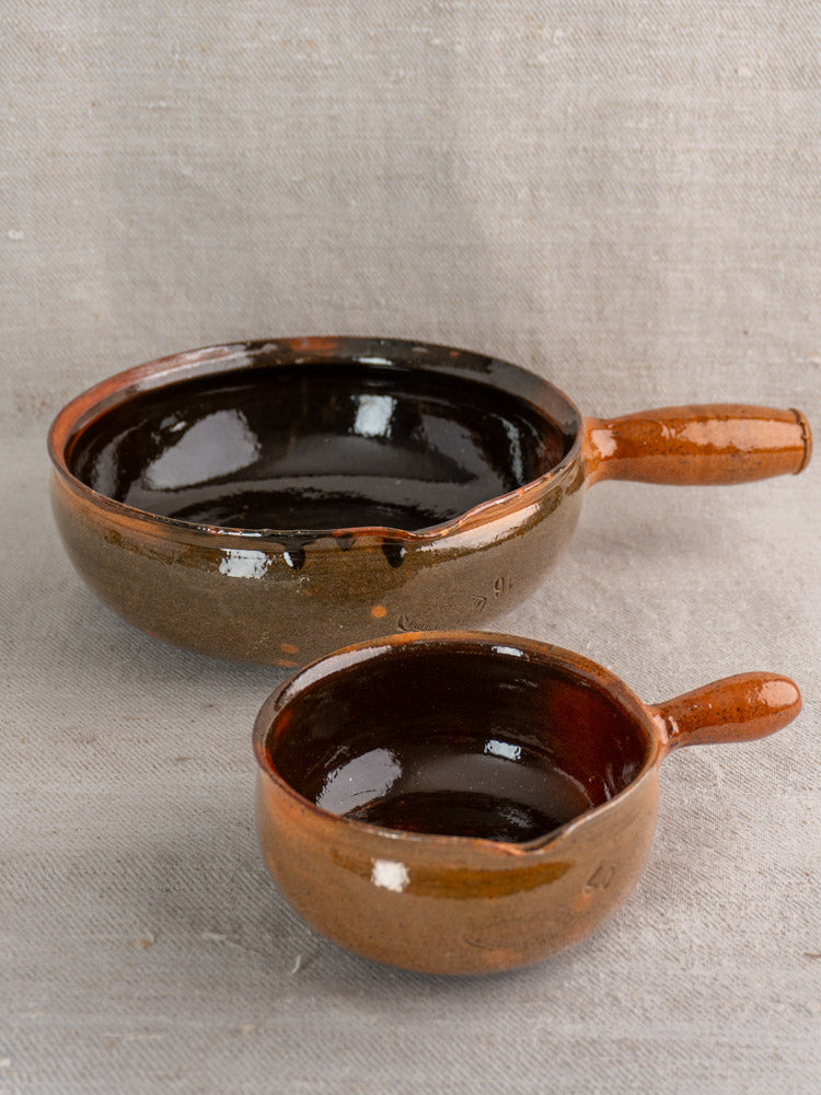 Pair of mid-century French Grandjean Jourdan Vallauris brown glazed ceramic poêlons with handles on light textured fabric.