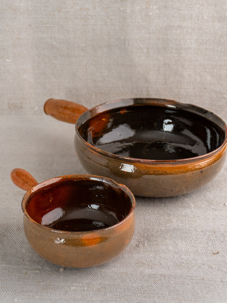 Pair of antique French Grandjean Jourdan Vallauris poêlon bowls with brown glazed handles on beige fabric.