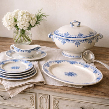 Vintage white china with blue floral motifs—plates, gravy boat, tureen, platter—on rustic wood table with white flowers.