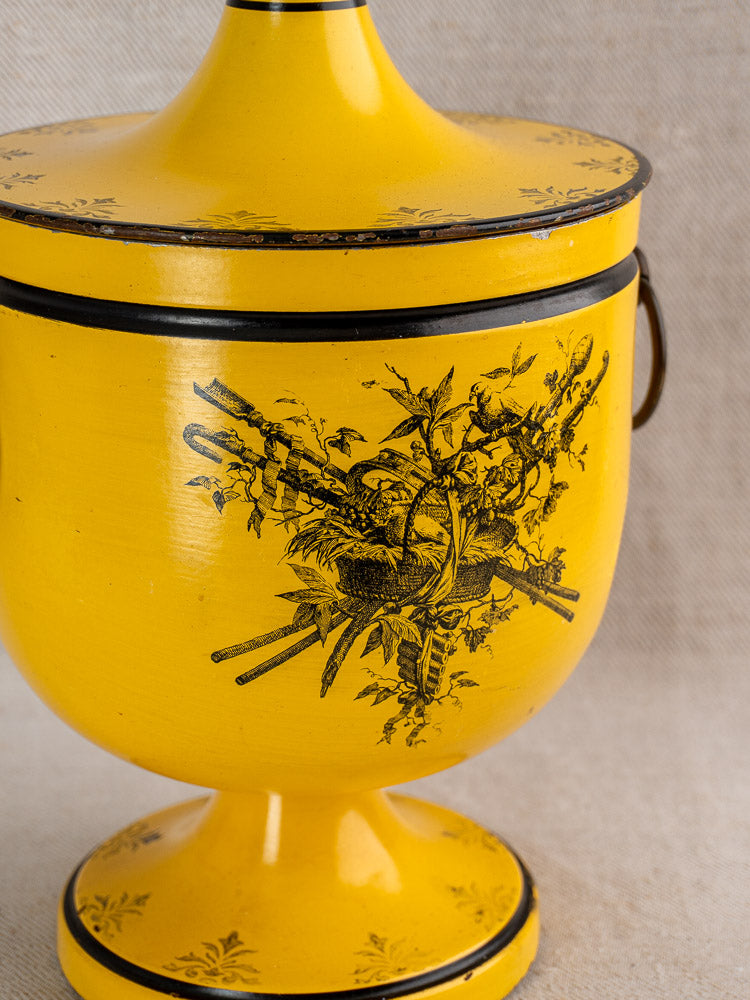 Yellow French Directoire style urn with black bird and branch motif, matching lid, on neutral fabric.