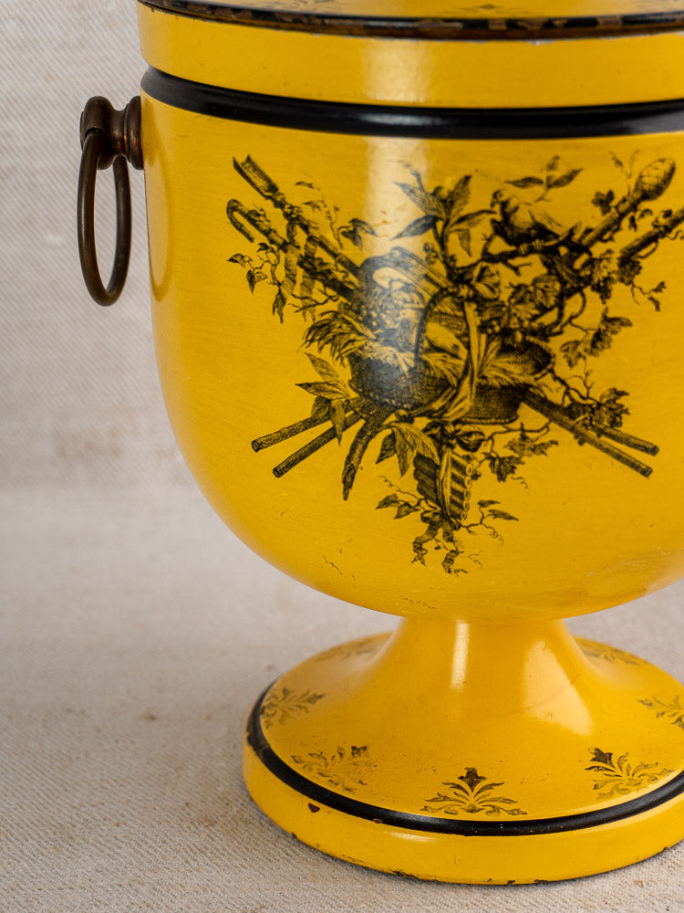 Vintage French Tôle Peinte champagne cooler in yellow Directoire style with black motifs and metal ring handle, on neutral fabric background.