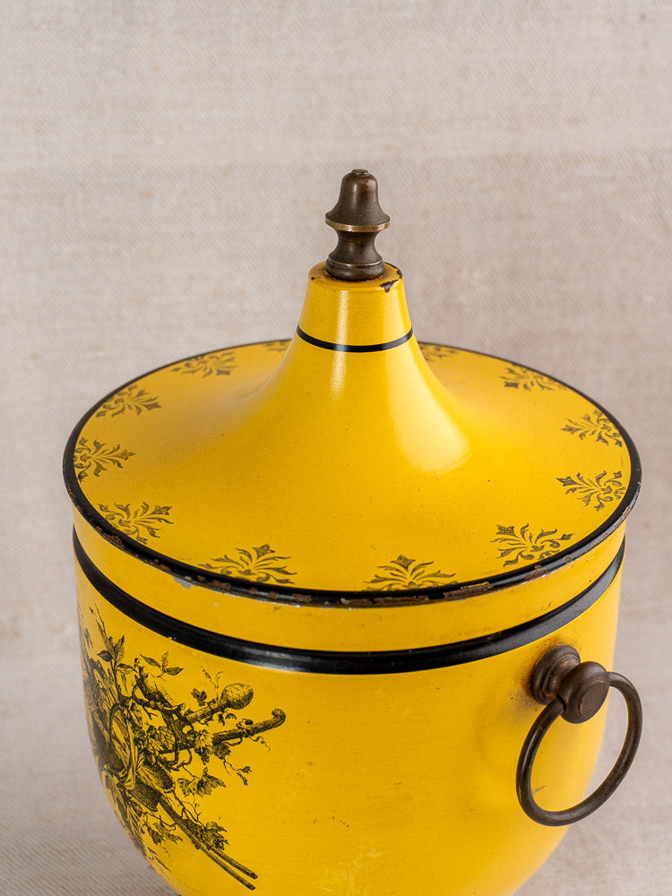 Vintage French yellow tôle peinte champagne cooler with pointed lid, black floral accents, and metal ring handle on beige fabric.