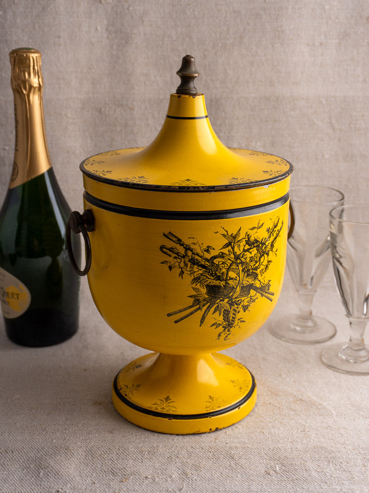 French yellow Tôle Peinte Directoire champagne cooler with black floral motifs and lid on table.