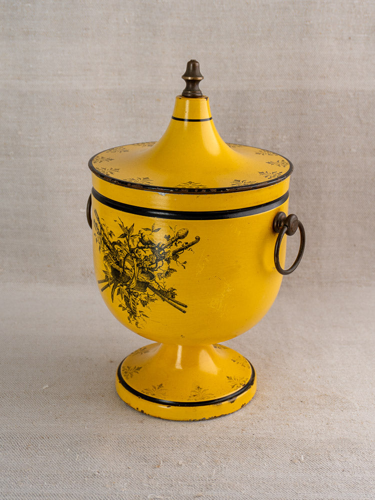 Vintage French Directoire-style tôle peinte champagne cooler with yellow finish, black floral motifs, and metal handles on light textured fabric.