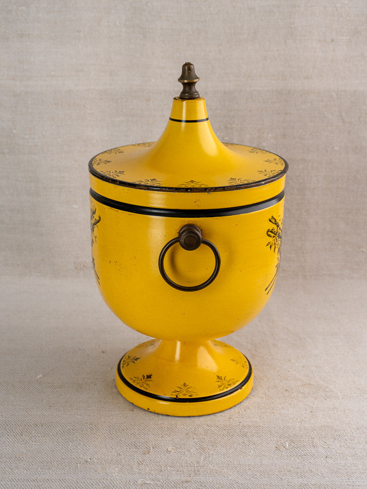 Vintage French yellow tôle peinte champagne cooler with lid, black trim, ornate motifs, ring handle; beige fabric backdrop.