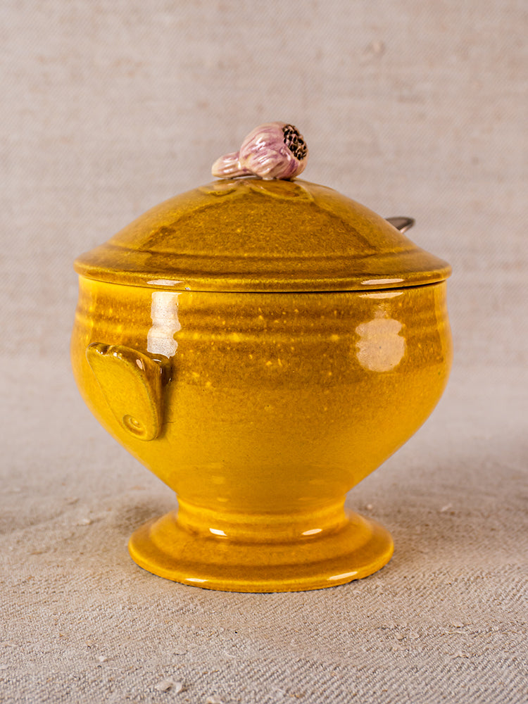 Yellow French ceramic soup tureen with pink garlic bulb handle, on a textured beige surface.