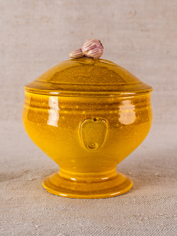 Glazed yellow French ceramic soupière with pink garlic-shaped handle, signed Eyguières, on beige surface.