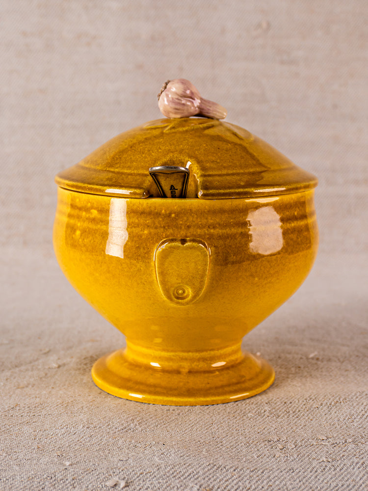 Yellow ceramic French soupière-style sugar bowl with lid, pink bird finial, spoon slot, on neutral textured surface.