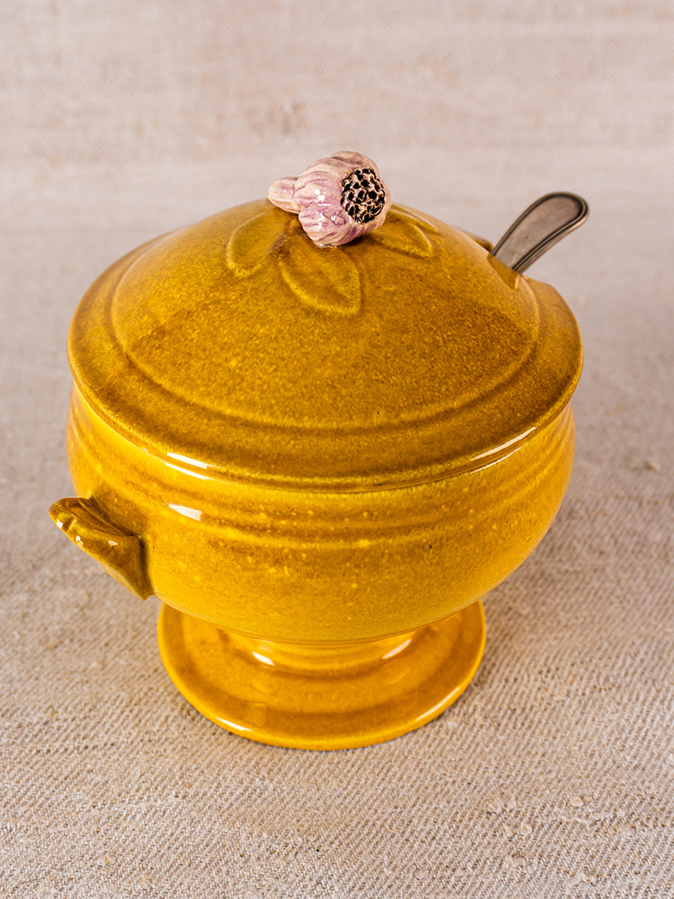 Yellow French ceramic soupière with garlic-shaped lid handle and spoon slot on beige textured surface.