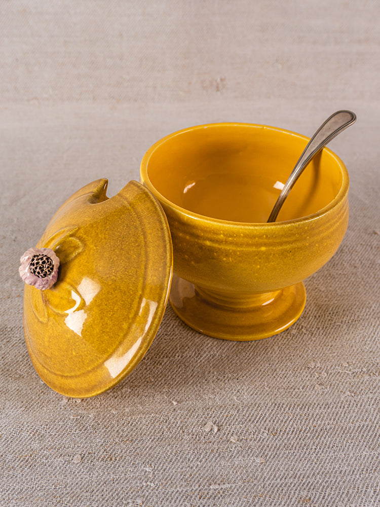 Vintage French yellow Eyguières Provençal tureen with matching lid, metal spoon, on beige textured fabric.