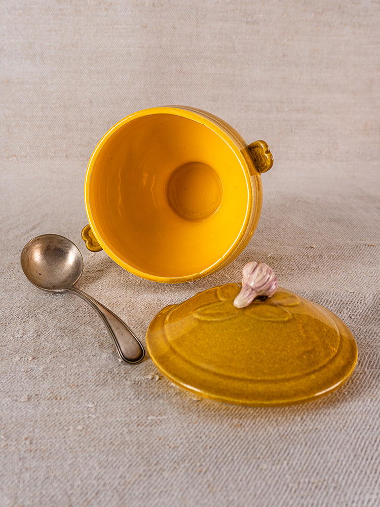 Vintage French yellow soupière, signed Eyguières Provençal tureen with garlic-shaped lid handle, open beside metal ladle on beige textured surface.