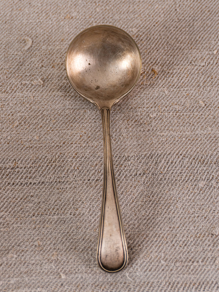 Vintage French yellow soupière signed “Eyguières” with antique silver spoon on textured beige fabric.