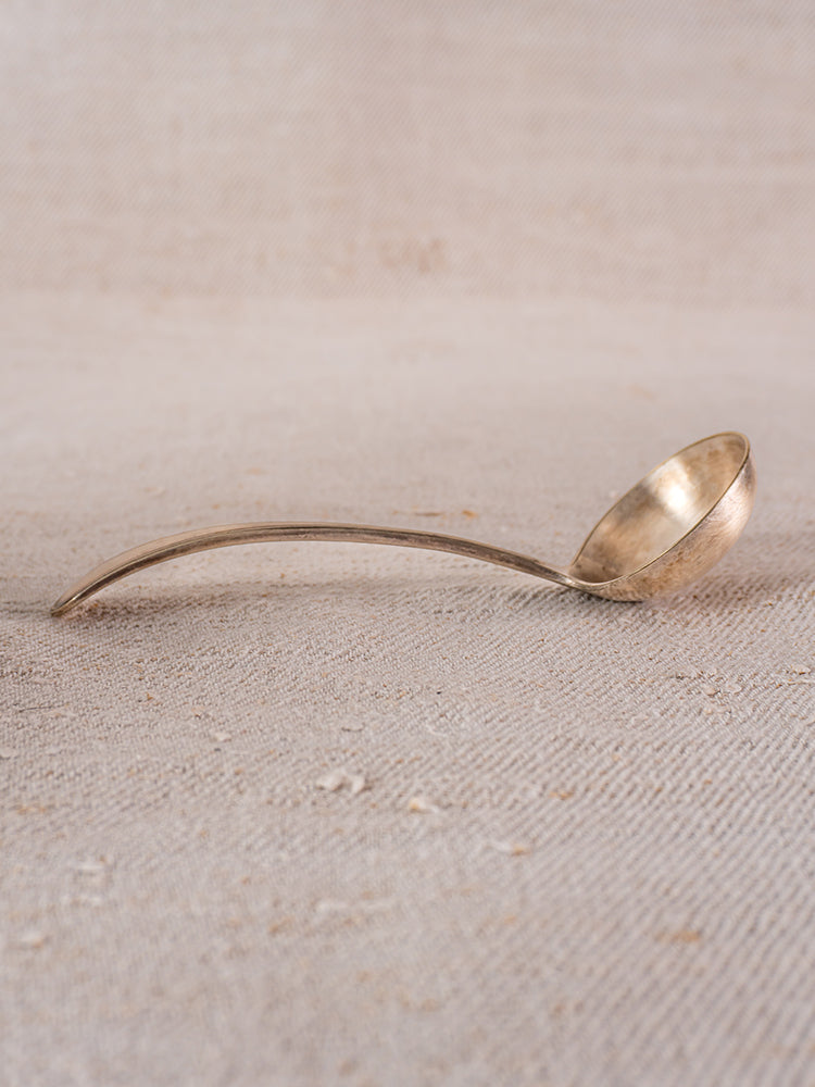 Gold-toned brass ladle with curved handle on beige textured surface, evoking Provence table settings.