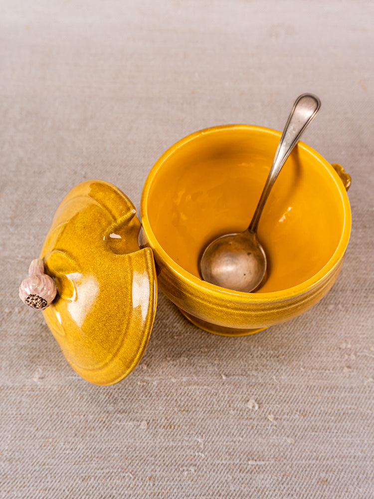Signed Eyguières French yellow soupière with lid and metal spoon on textured beige surface.
