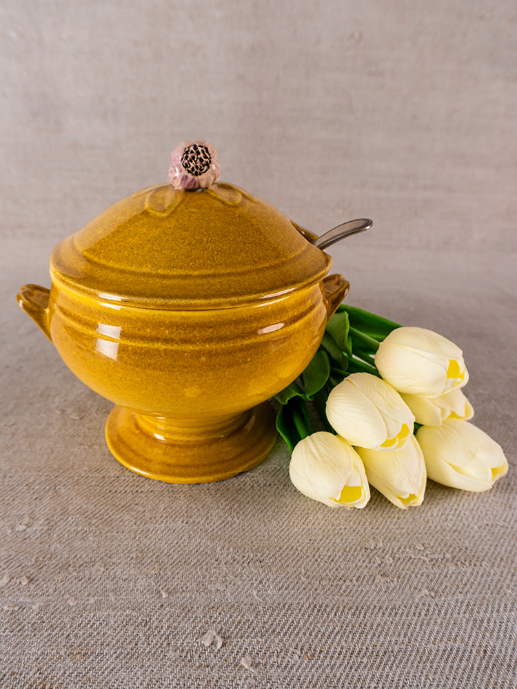 Vintage French yellow Eyguières Provençal tureen with lid and spoon, beside white tulips on beige fabric.