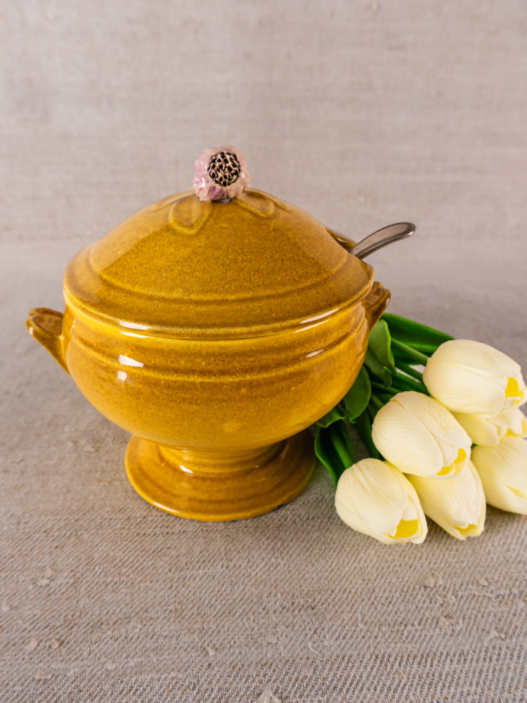 Signed Eyguières French yellow soupière with lid and ladle beside white tulips on neutral surface.