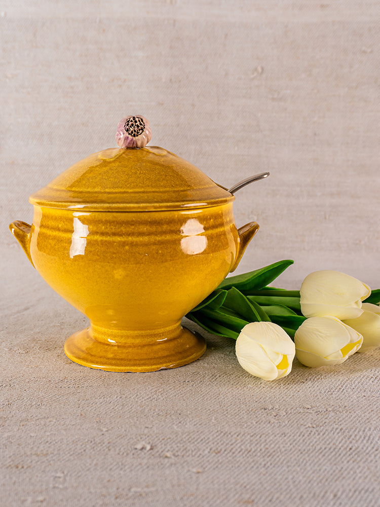 Signed Eyguières Provençal yellow soupière with lid and spoon beside white tulips on fabric.