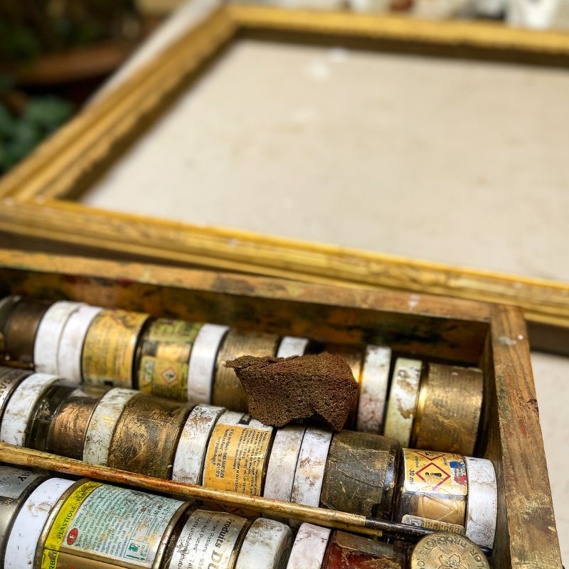 Artist’s paint box with assorted well-used paint tubes and sponge, blank canvas and wooden frame in background outdoors.